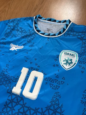 Israel fotbollströja  - Säljer en blå Israel fotbollströja från Reebok med nummer 10 och B. NETANYAHU på ryggen. Tröjan har geometriskt mönster i svart, vit krage med blå detaljer och klubbmärke på bröstet. Perfekt för dig som gillar fotboll och vill sticka ut på planen och perfekt inför sommaren.                                         Skriv om ni har några funderingar!😃