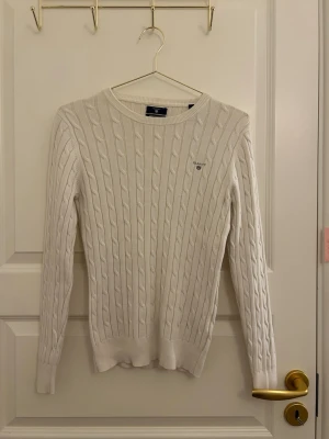 Vit kabelstickad tröja från Gant - Kabelstickad vit tröja från Gant med rund halsringning och diskret logga broderad på bröstet. Tröjan har långa ärmar och ribbade muddar vid ärmslut och nederkant. Klassisk och stilren design som passar till mycket.