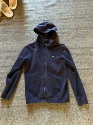 Mörkblå zip hoodie från Lyle & Scott - Snygg mörkblå hoodie från Lyle & Scott Junior med dragkedja framtill och klassisk gul logga på bröstet. Hoodien har känguruficka och ribbade muddar vid ärmslut och nederkant. Perfekt för en chill och stilren look. Storlek 170/176 de vill säga 15/16år