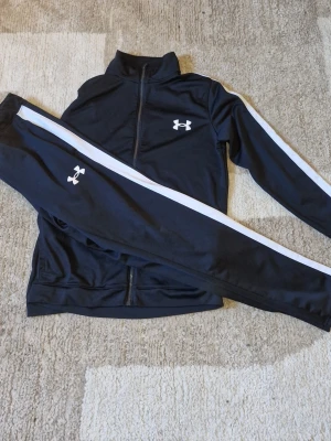 Svart träningsset från Under Armour - Snyggt svart träningsset från Under Armour med vita ränder längs ärmar och ben. Setet består av en zip-jacka och matchande byxor, båda med logga på bröst och lår. Tillverkat i ett mjukt och stretchigt funktionsmaterial som passar perfekt till träning. Köpte detta sättet endast för träning, men det har inte blivit av något och jag har inte ens använt plaggen ordentligt en enda gång, endast testat det! Inte förhandlingsbart pris, ett rimligt pris för ett helt orört träningsställ. 
