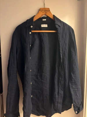 Mörkblå linneskjorta These Glory Days - Stilren mörkblå skjorta från These Glory Days i slim fit-modell. Skjortan är tillverkad i linne och har klassisk krage, långa ärmar och vita knappar framtill. Perfekt för en avslappnad men ändå snygg look.