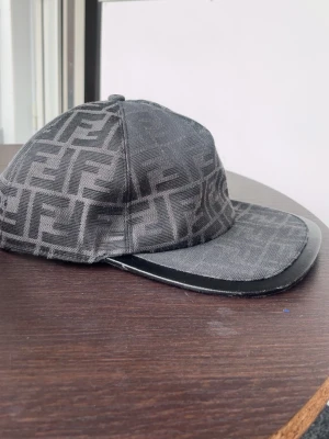 Svart keps med Fendi-logga - Säljer en svart keps från Fendi med det klassiska FF-monogrammet över hela kepsen. Kepsen har böjd skärm med en snygg läderdetalj längs kanten och är tillverkad i ett tygmaterial med insida i svart textil. Perfekt för dig som gillar exklusiva accessoarer.