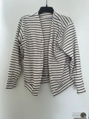 Randig kofta från FU - Säljer en vit kofta med svarta horisontella ränder från FU. Koftan har knappar framtill och är tillverkad i ett mjukt, tunt material. Perfekt för lager på lager och enkel att matcha med olika outfits. Långärmad och klassisk design.