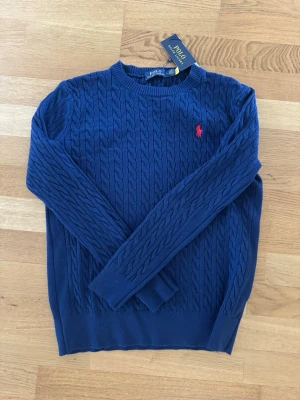 Marinblå kabelstickad tröja Polo Ralph Lauren - Kabelstickad marinblå tröja från Polo Ralph Lauren med rund halsringning och röd broderad logga på bröstet. Storlek medium men passar en small pågrund av att den är liten i storleken. 