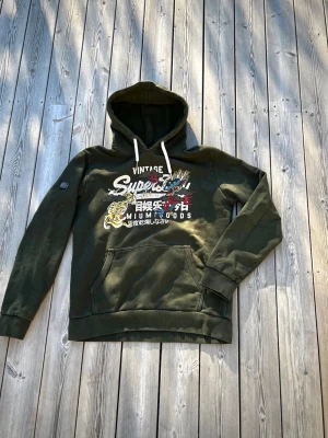 Superdry hoodie - En riktigt fet Superdry hoodie med en ovanligt och snyggt tryck på bröstet. Den har en fräsch och snygg grön färg som inte sticker ut för mycket och inga defekter förutom en slitning som inte syns när man har på sig den. Den är storleken Xl men passar mer som L vilket är vanlig i vintage kläder. Kom pm för frågor 