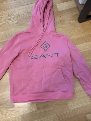 Rosa hoodie från GANT med logga - Snygg rosa hoodie från GANT med stor logga och emblem på bröstet. Klassisk känguruficka framtill, ribbade muddar och justerbar huva med dragsko. Perfekt för en avslappnad och trendig look. Tillverkad i mjukt material för extra komfort.