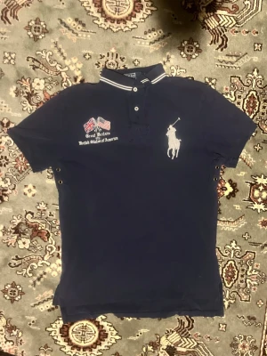 Vintage Marinblå pikétröja från Polo Ralph Lauren - Snygg marinblå pikétröja från Polo Ralph Lauren med vit broderad logga på bröstet och detaljerade flaggor. Tröjan har korta ärmar, knappar vid kragen och vita ränder på kragen. Perfekt för en sportig och stilren look.
