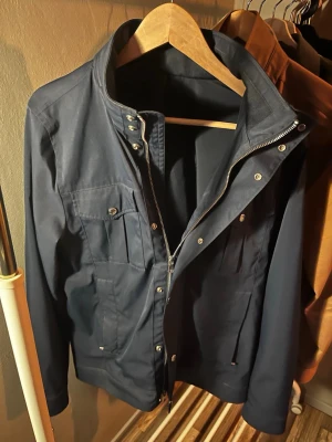 Mörkblå vindjacka från J. Lindeberg - Stilren mörkblå vindjacka från J. Lindeberg med hög krage, dragkedja och tryckknappar framtill. Jackan har fyra fickor med lock och knapp, samt justerbara ärmslut. Tillverkad i ett lätt och slitstarkt material som passar perfekt till vardagslooken. I storlek S