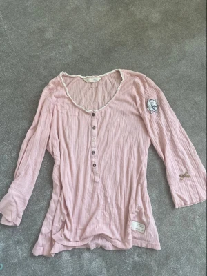 Ljusrosa 2000s topp  - Supersöt ljusrosa blus från Odd Molly med trekvartsärm, knappar framtill och spetskant vid halsen. Blusen har en broderad detalj på ärmen och en liten rosett. Perfekt för dig som gillar romantisk stil.