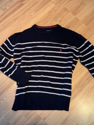 Marinblå randig tröja Polo Ralph Lauren - Stickad marinblå tröja från Polo Ralph Lauren med vita horisontella ränder och röd broderad logga på bröstet. Tröjan har rund halsringning med en röd detalj på insidan och är tillverkad i mjukt material. Perfekt för en klassisk och avslappnad stil. Ny, kvitto finns möjligen på mail om jag kan hitta det. 