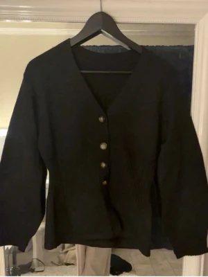 Svart kofta med knappar - Stilren svart kofta med v-ringning och fyra knappar framtill. Koftan har ribbade detaljer vid midjan och långa ärmar. Perfekt att slänga på sig över en t-shirt eller linne för en snygg lager-på-lager-look.