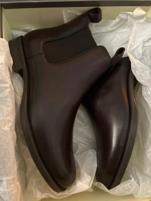 Bruna Chelsea boots från Zenobi - Snygga bruna Chelsea boots från Zenobi i slätt läder med elastiska sidpaneler och dragflik bak. Klassisk och stilren design med låg klack och rundad tå. Yttersulan är mönstrad för extra grepp och bootsen är tillverkade i Italien.
