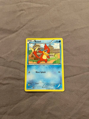 Pokémonkort Buizel Wave Splash - Pokémonkort med Buizel-motiv från Pokémon-serien. Kortet har en blå och gul ram, med en tecknad Buizel i mitten och texten 'Wave Splash'. Materialet är glansigt papper/kartong.