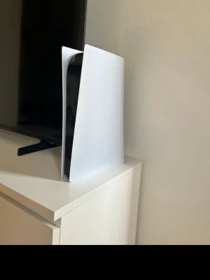 PlayStation 5 spelkonsol - Säljer en PlayStation 5 från Sony. Konsolen har en futuristisk design med vita, böjda sidopaneler och svart mittdel. Perfekt för gaming och streaming, med kraftfull prestanda och stöd för 4K-upplösning.