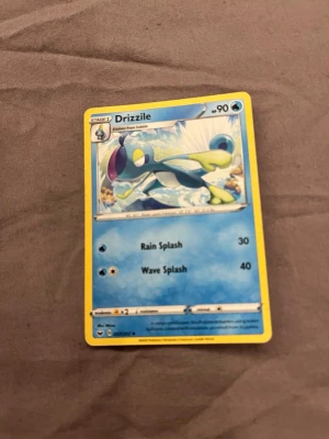 Pokémonkort Drizzile - Säljer ett Pokémonkort med Drizzile, en vattenbaserad Pokémon. Kortet har blå och gula färger med en bild på Drizzile i vattenmiljö. Kortet har attacker som Rain Splash och Wave Splash och är inramat med gul kant. Perfekt för samlare eller Pokémonfans.
