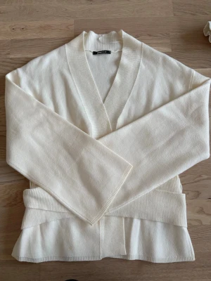 Krämvit kofta från Gina Tricot - Säljer en krämvit kofta från Gina Tricot med breda ribbade muddar och öppen front. Koftan har en avslappnad passform och är tillverkad i mjukt stickat material. Helt oanvänd.