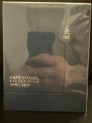 Café Citadel Eau de Parfum 100ml - Café Citadel Eau de Parfum från Aurora Scents kommer i en elegant, mörkgrå rektangulär förpackning med guldfärgad text. Flaskan rymmer 100 ml och designen ger en lyxig och modern känsla. Perfekt för dig som gillar stilrena och exklusiva dofter.