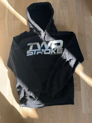 Svart TWO STROKE hoodie - Svart hoodie från TWO STROKE med stor vit logga på bröstet och en stor grafisk print på ryggen med en hand som visar långfingret i en färgglad ruta. Klassisk huva med dragsko och känguruficka framtill. Perfekt för dig som vill sticka ut och gillar streetwear.