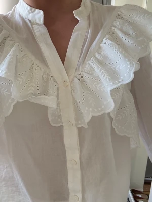 Vit blus med broderade volanger - Säljer en vit blus med knappar framtill och stora broderade volanger över bröstet och axlarna. 