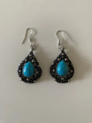 Svart-silver örhängen med turkos sten - Coola droppformade örhängen i svart och silver med en stor turkos sten i mitten. Dekorerade med små pärlor och detaljer runt om stenen. Hängande modell med krok. Perfekta för att ge din look en bohemisk touch.