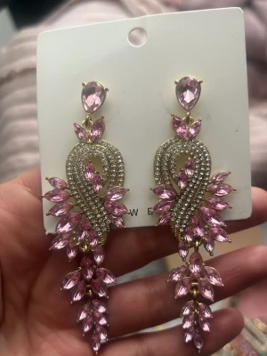 Långa örhängen med rosa stenar - Statementörhängen i guldton med massor av gnistrande rosa stenar i olika former. Örhängena har en böjd design med små klara stenar och droppformade rosa kristaller som ger en lyxig och iögonfallande look.