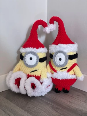 Virkade Minion-julfigurer - Två handgjorda virkade Minion-figurer klädda i tomtedräkt med röda och vita detaljer, grå glasögon och svarta skor. Perfekta som juldekoration eller present till någon som gillar Minions och julpynt. Säljes som ett par.