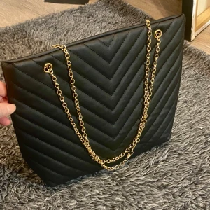 Större. Axelväska  med randigt mönster  - Större axelväska svart i konstläder med randigt mönster och en kedja i Ljusguld som axelband , knapplåset och dragkedja inuti 