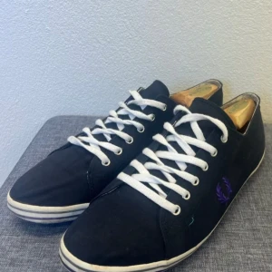 Marinblåa Fred Perry sneakers strl 45 - Klassiska marinblåa sneakers från Fred Perry med vit sula och vita snören. Skorna har en låg profil och är tillverkade i textil med diskret lila Fred Perry-logga på sidan. Perfekta för en clean och stilren look.