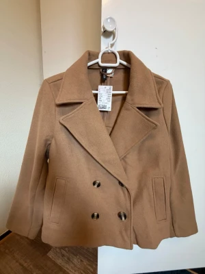Kort kamelfärgad trenchcoat H&M - Helt ny och stilren kort dubbelknäppning trenchcoat i kamelfärg från H&M. Jackan har två fickor framtill och är tillverkad i ett mjukt material som ger en klassisk look. Perfekt för dig som gillar minimalistisk och trendig stil. Passar utmärkt till våren eller hösten. Jackan är slutsåld i butik och säljs inte online. 