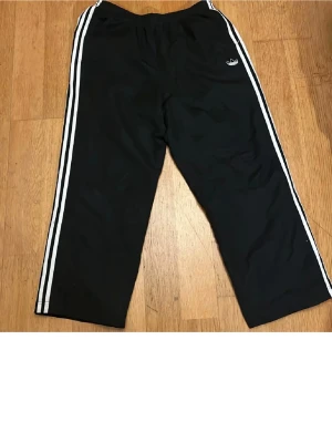 Svarta Adidas träningsbyxor - Svarta träningsbyxor från Adidas med klassiska vita ränder längs sidorna och liten Adidas-logga framtill. Byxorna har rak passform och resår i midjan. Perfekta för sport eller chill.