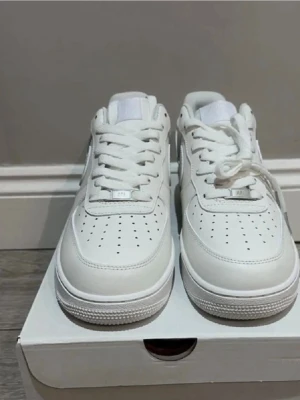 Vita Nike Air Force 1 sneakers - Klassiska helvita Nike Air Force 1 sneakers med lågt skaft. Skorna har en chunky sula, perforerad tåbox och är tillverkade i läder med textildetaljer. Perfekta för en clean och stilren look. Helt nya 