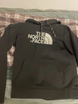 Svart hoodie från The North Face - Snygg svart hoodie från The North Face med stor vit logga på bröstet. Klassisk modell med huva, dragsnören och stor magficka. Perfekt för en avslappnad streetstyle-look och riktigt skön att bära.