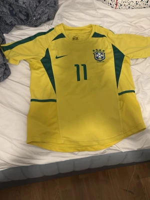 Brasilien tröja  - Gul Brasilien fotbollströja från Nike med gröna detaljer, nummer 11 på bröstet och CBF-emblem. Klassisk rund hals och korta ärmar, tillverkad i lätt och ventilerande material som passar perfekt till träning eller match.