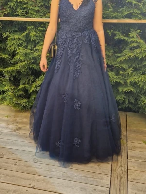 Marinblå långklänning med spetsdetaljer - Elegant marinblå långklänning med v-ringning och vackra spetsapplikationer. Klänningen har snörning i ryggen och är ärmlös med tunna axelband. Perfekt för bal eller festliga tillfällen.