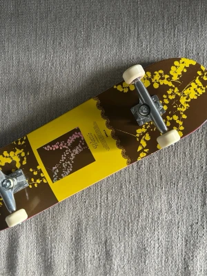 Impala skateboard - Helt oanvänd 8,5tum Impala skateboard med unik design i gult och brunt, dekorerad med blommönster och grafiskt tryck på ovansidan. Brädan har vita hjul och silverfärgade trucks. Materialet är trä med tryckt motiv och klassisk form.