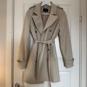 Beige lyxig trenchcoat  - Snygg beige trenchcoat från storlek 34 med klassisk design. Jackan har knappar och ett bälte i midjan för en elegant look. Perfekt för vår och höst med sina långa ärmar och stilrena detaljer. Från MQ