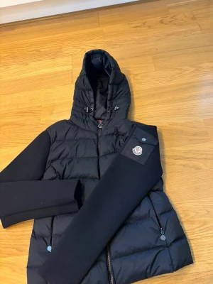 Svart cardigan från Moncler - Svart pufferjacka från Moncler med quiltad kropp, släta ärmar och huva med dragsko. Jackan har dragkedja framtill, två fickor med dragkedja och Moncler-logga på ena ärmen. Perfekt för vintern och riktigt snygg streetstil.