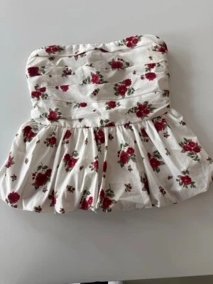 Blommig tubtopp från Hollister - Supersöt vit tubtopp från Hollister med röda blommor och smockad rygg. Toppen har en volangig nederdel och är perfekt för varma dagar. Den är axelbandslös och har en romantisk vibe som passar till jeans eller kjol.