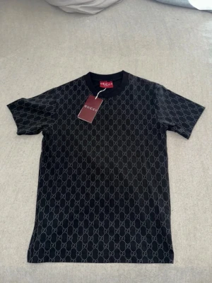 Svart Gucci t-shirt med monogram - Snygg svart t-shirt från Gucci med klassiskt GG-monogram över hela tröjan. T-shirten har rund halsringning och korta ärmar. Perfekt för dig som vill ha en stilren och lyxig look med tydlig logga.