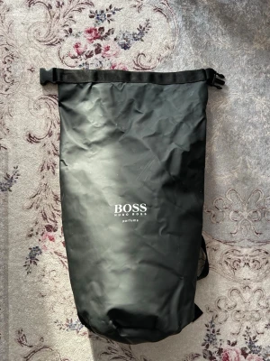 Svart ryggsäck från Hugo Boss - Cool svart ryggsäck från Hugo Boss med rullstängning och justerbara axelband med logga. Tillverkad i vattenavvisande material, perfekt för att bära med sig grejer när det regnar. Minimalistisk design med diskret tryck framtill.