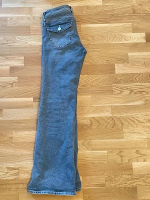 Blå bootcut jeans med fickdetaljer - Säljer ett par klassiska blå bootcut jeans med snygga fickor bak som har lock och knapp. Jeansen har en lätt utsvängd passform nertill och är i ett mjukt denimtyg. Perfekta för dig som gillar retrostil och vill ha något bekvämt och stilrent.