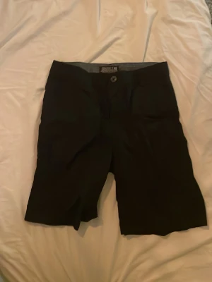 Svarta shorts från ORGNLS - Svarta shorts från ORGNLS med klassisk passform och knappstängning framtill. Perfekta för sommaren med enkel design och bekväm midja. Tillverkade i ett mjukt material som känns skönt mot huden.