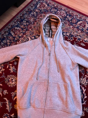 Grå zip-hoodie från Burberry - Säljer en grå hoodie från Burberry med dragkedja och stora fickor framtill. Huvan har klassiskt Burberry-mönster på insidan och snören med metalländar. Perfekt för dig som vill ha en stilren och exklusiv look.