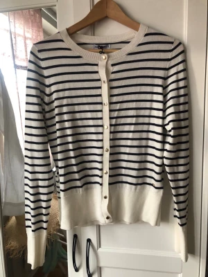 Vit- & blårandig kofta från Holly & Whyte Stl S - Säljer en klassisk vit kofta från Holly & Whyte  med marinblå ränder och rund hals. Koftan har långa ärmar och guldfärgade knappar framtill. Perfekt för dig som gillar en tidlös och stilren look.