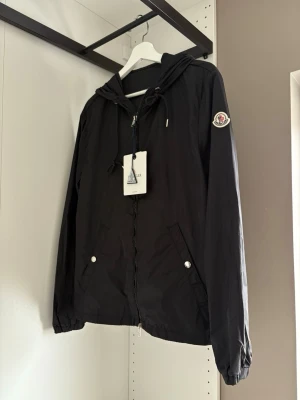 Svart Moncler vindjacka med huva - Svart vindjacka från Moncler med huva och dragsko, två snedställda fickor med silverfärgade knappar och Moncler-logga på ärmen. Jackan har en diskret röd och vit detalj på huvan och är tillverkad i lätt, vattenavvisande material.