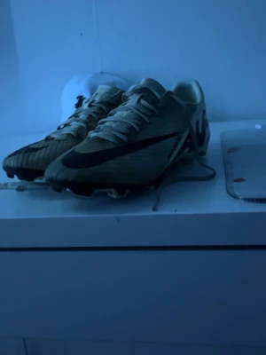 Nike Mercurial fotbollsskor beige - Säljer ett par Nike Mercurial fotbollsskor i beige med svart swoosh och detaljer. Skorna har en smal och aerodynamisk form, tillverkade i syntetmaterial för bra grepp och komfort på planen. Perfekta för dig som vill ha snabbhet och kontroll.