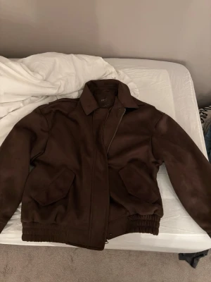 Mörkbrun mocka med krage - Snygg mörkbrun mocka med klassisk krage och två stora fickor framtill. Jackan har resår vid midjan och ärmarna för en schysst passform. Perfekt för dig som vill ha en stilren och tidlös look.