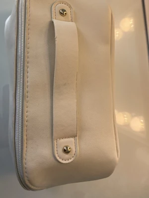 Beige necessär med handtag - Necessär i vit färg med stilrent handtag och dragkedja. Insidan har ett extra fack med dragkedja för småprylar och en vägg i mitten vilket delar necessären i två separata fack. 