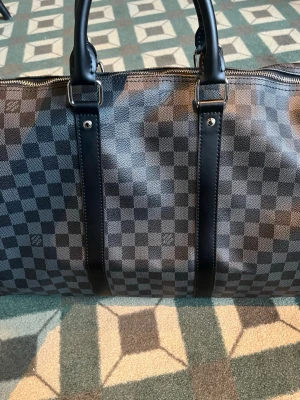 Louis Vuitton Damier Graphite resväska - Snygg och rymlig resväska från Louis Vuitton i det klassiska Damier Graphite-mönstret. Väskan är svart och grå med rutigt mönster, har dubbla handtag och detaljer i svart skinn. Perfekt för weekendresor och har en lyxig känsla.