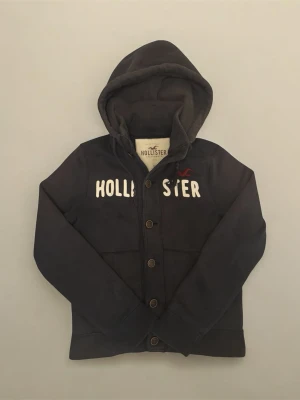 Svart vintage cardigan med knappar från Hollister - Säljer en snygg svart vintage cardigan från hollister i storlek Xl, men passar L (180-190cm). Tröjan är i ganska bra skick 7/10. Tröjan är också väldigt sällsynt och eftertraktad då den inte tillverkas längre. Priset är ej hugget i sten så kom gärna med bud!